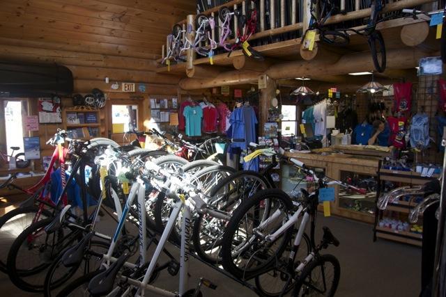 Interior of a bicycle shop featuring a variety of bikes in the foreground, with colorful clothing and accessories displayed on racks and shelves. The shop has a wooden decor, creating a warm atmosphere, and a counter visible in the background.