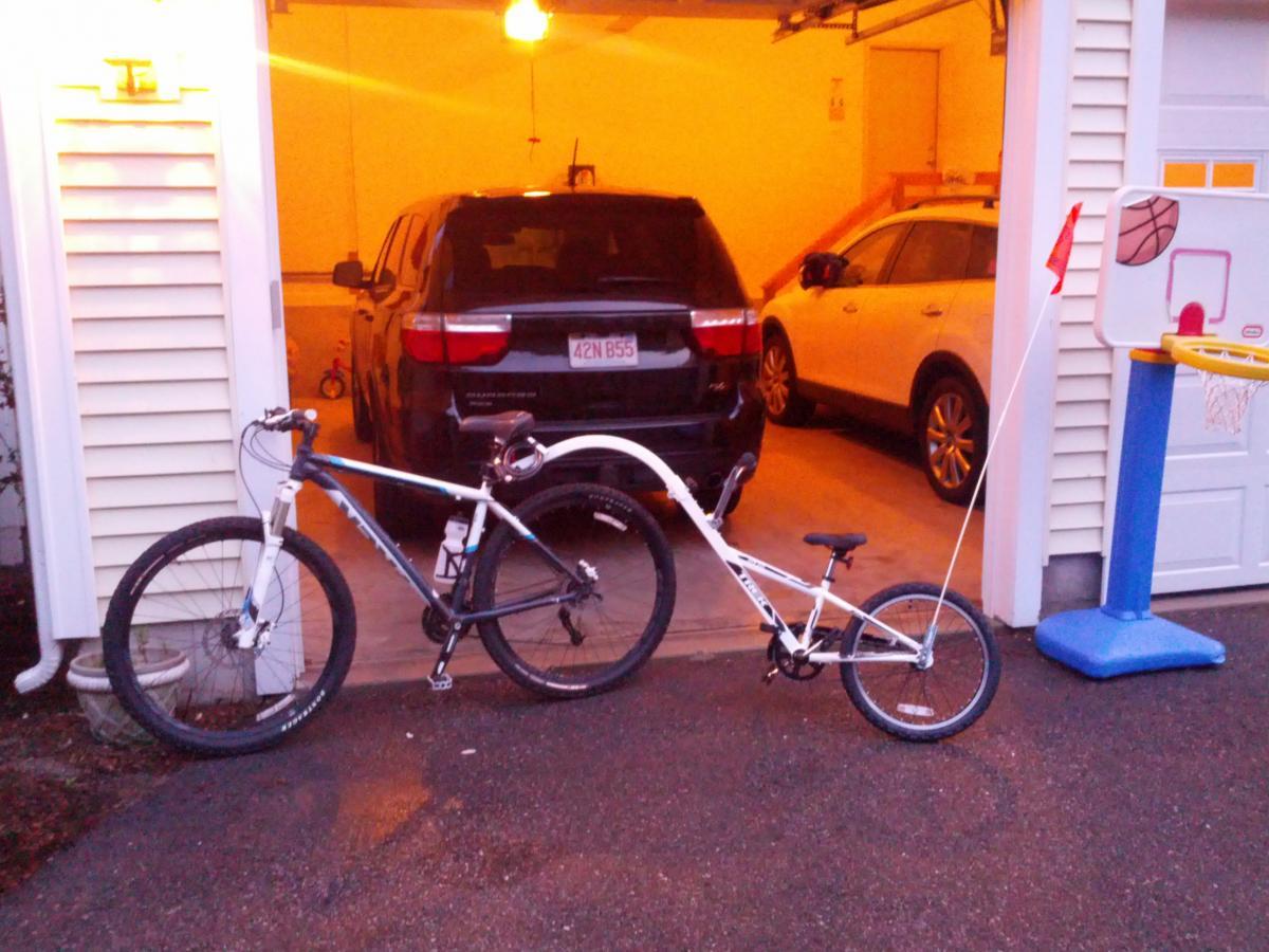 Trek X-Caliber: A mountain bike parked outside a garage, attached to a smaller bike with a tow bar. The garage contains two vehicles, and a small basketball hoop is visible nearby. The scene is illuminated by warm light from the garage.