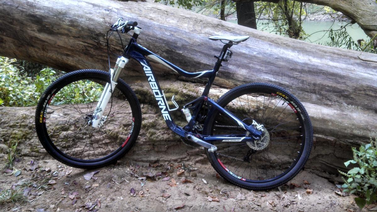 Airborne Hobgoblin: A mountain bike leaning against a large fallen log in a wooded area, surrounded by greenery and dirt. The bike features a blue and white frame with the word "AIRBORNE" prominently displayed. The terrain appears sandy with scattered leaves.