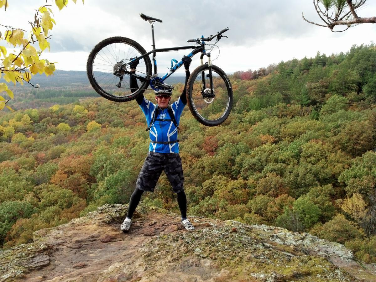 A cyclist in a blue jersey and helmet stands triumphantly on a rocky outcrop, holding a mountain bike above their head. They are surrounded by a vibrant landscape of autumn foliage, featuring shades of green, orange, and yellow, under a cloudy sky. Clarence mountain bike trail.