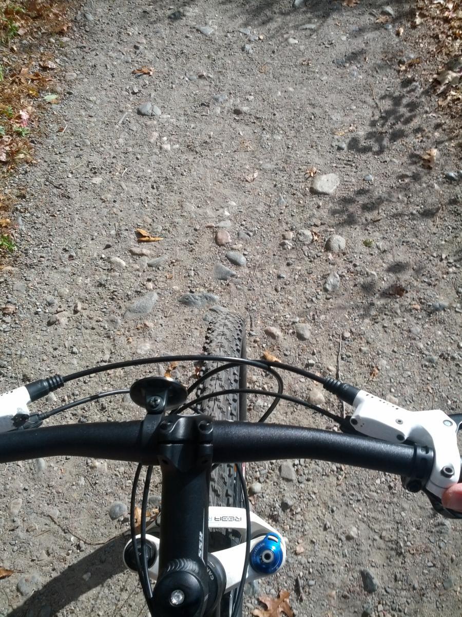 A vantage point from the handlebars of a mountain bike, showing a gravel path ahead. The ground is scattered with small pebbles and dried leaves, indicating an outdoor trail surrounded by nature. The bike's handlebars and controls are visible in the foreground. Cutler Park mountain bike trail.