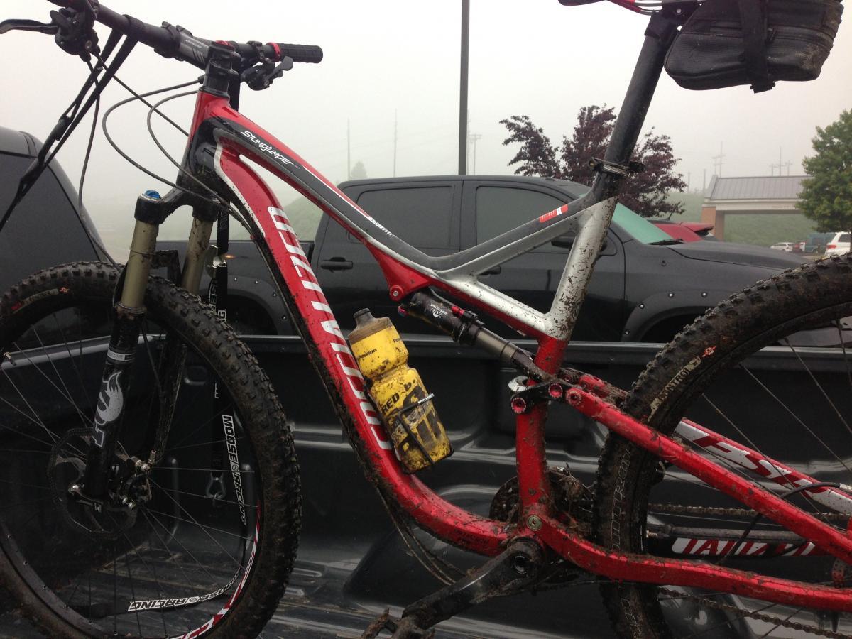 Specialized Stumpjumper FSR Comp 29: Mountain bike resting in the bed of a pickup truck, showing signs of mud and dirt, indicating recent use. A yellow water bottle is attached to the frame, and the background features a misty atmosphere with parked vehicles.