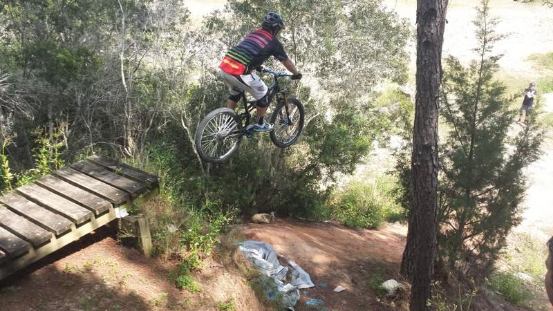 A mountain biker in mid-air performing a jump off a wooden ramp, surrounded by trees and vegetation. The rider is wearing a helmet and colorful clothing, with a trail visible in the background. Santos mountain bike trail.