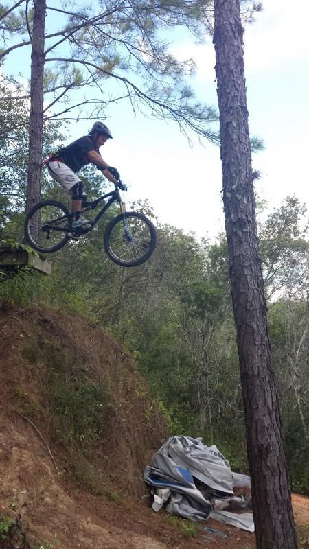 A mountain biker mid-jump off a dirt ramp, with trees in the background and a hillside covered in greenery. The cyclist is wearing a helmet and protective gear, and there is a tarp-covered pile on the ground below. Santos mountain bike trail.