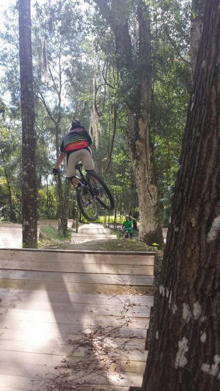 A cyclist performing a jump on a dirt bike track surrounded by trees. The rider is airborne with their bike elevated above a wooden ramp, showcasing an action-packed moment in a natural setting. In the background, another person is visible, also riding a bike on the track. Santos mountain bike trail.