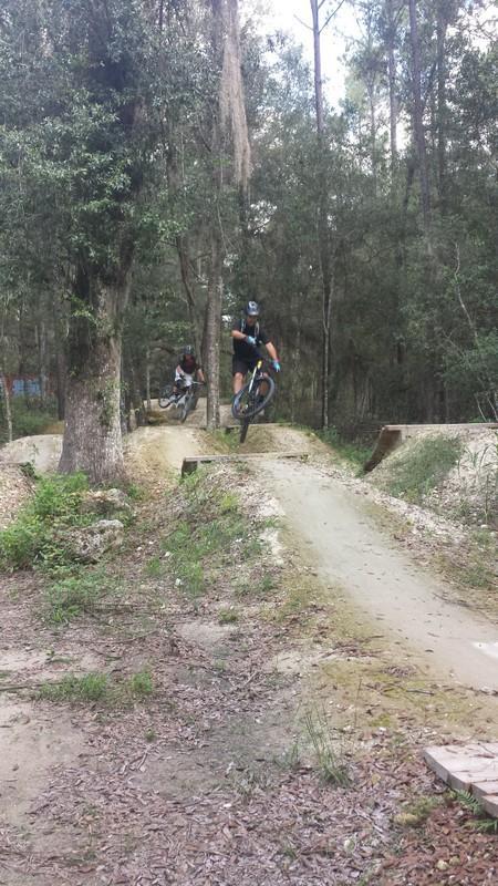 Two mountain bikers performing jumps on a dirt trail in a wooded area. The scene shows one rider in the foreground airborne above a jump, while another rider follows closely behind on a parallel path. Surrounding trees and underbrush create a natural environment, with soft dirt paths and small jumps visible in the landscape. Santos mountain bike trail.