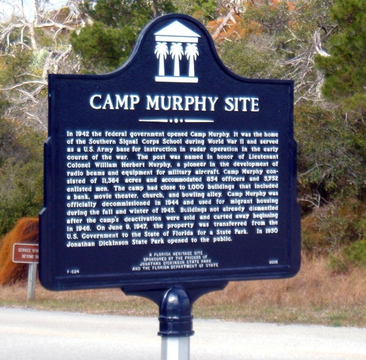Black historical marker for Camp Murphy Site, detailing its establishment in 1942 as a U.S. Army base for radar instruction during World War II. The text includes information about its size, facilities, and decommissioning in 1944, along with its subsequent use and transfer to the state in 1947. Palm tree motifs are featured at the top. Jonathan Dickinson State Park mountain bike trail.