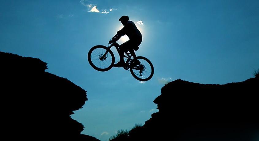 A silhouetted mountain biker jumping across a canyon, with the sun shining behind them against a blue sky. Wilkins Peak Trails mountain bike trail.