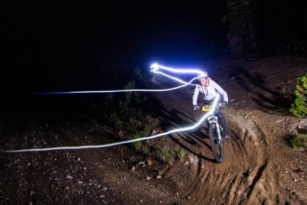 A mountain biker riding through a dirt trail at night, illuminated by bike lights that create dynamic light trails in the air. The rider is wearing a helmet and a light-colored outfit, navigating a curved path surrounded by trees. The scene captures the excitement and energy of night biking.
