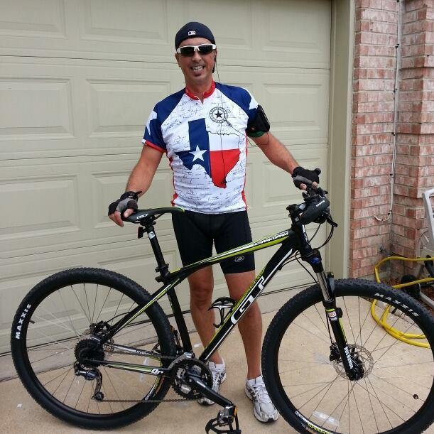 GT Karakoram: A man wearing a Texas-themed cycling jersey stands beside his mountain bike, smiling in front of a garage door. He is wearing cycling shorts and gloves, with sunglasses and a cap. The bike features thick tires and is parked on a driveway.