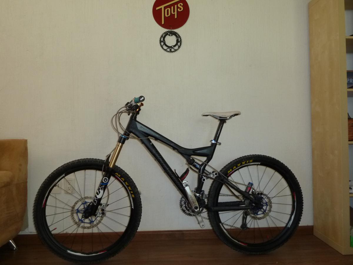Specialized Stumpjumper FSR Pro: A black mountain bike with a sturdy frame and thick tires is positioned against a light-colored wall, next to a brown couch and a wooden bookshelf. A circular red sign with the word "Toys" is mounted on the wall above the bike.