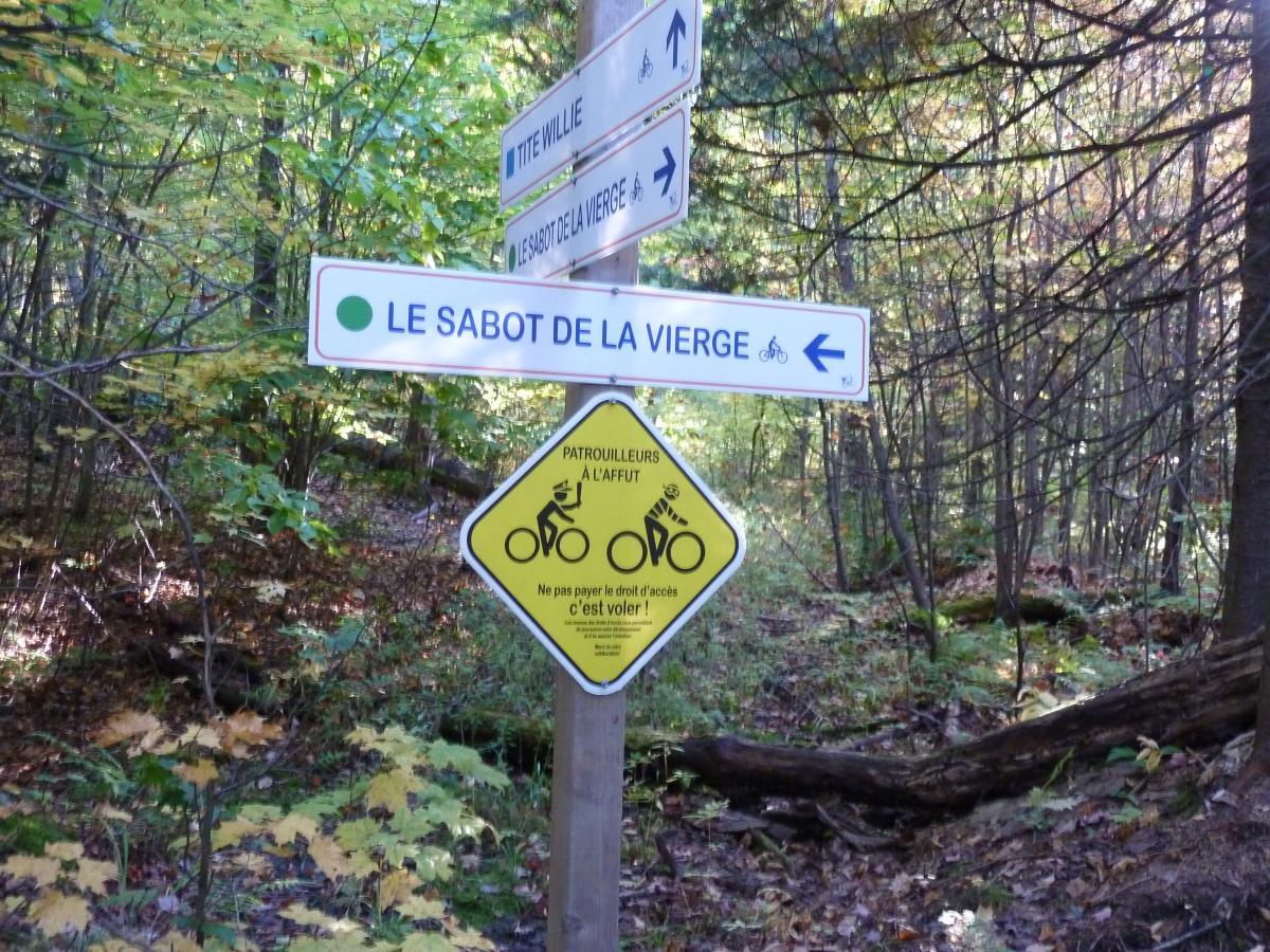 A wooden signpost in a wooded area, featuring directions for hiking and biking trails. The main sign points to "Le Sabot de la Vierge" with a green circle and an arrow to the left. Beneath, there is a yellow warning sign indicating bike patrols, along with a message in French advising not to pay for access. The background is filled with trees and autumn leaves. Tite Willie mountain bike trail.