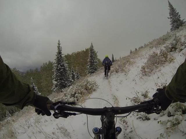 Mountain biking on a snowy trail, with a view from the rider's perspective showing a handlebars close-up and another cyclist ahead navigating through a winter landscape with snow-covered ground and evergreen trees. Scott's Bypass mountain bike trail.