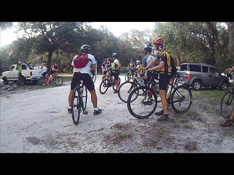 A group of cyclists gathered in a natural outdoor setting, preparing for a ride. Some cyclists are mounted on their bicycles while others are chatting or adjusting their gear. In the background, there are parked vehicles and trees, indicating a recreational area. The scene captures the camaraderie and enthusiasm of the biking community. Carlton Preserve mountain bike trail.