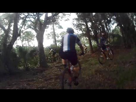 A group of mountain bikers riding along a trail surrounded by trees and lush greenery. The scene captures the dynamic nature of the sport, showcasing individuals in cycling gear navigating through a wooded area. Carlton Preserve mountain bike trail.