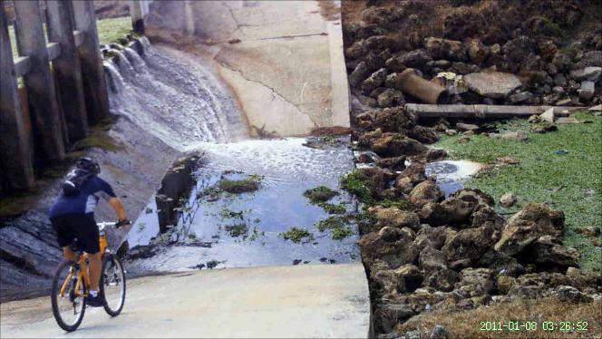A cyclist riding along a drainage channel with water flowing down the slope. The scene includes rocky terrain on one side and patches of greenery near the water. The image captures the cyclist positioned on the left, alongside a partially visible concrete structure and flowing water. North Port Mountain Bike Trails mountain bike trail.
