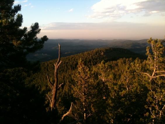 A scenic view of rolling hills and mountains, covered in dense forests, illuminated by warm sunlight. The sky above is partly cloudy, suggesting a tranquil evening atmosphere in a natural landscape. Bergen Peak mountain bike trail.