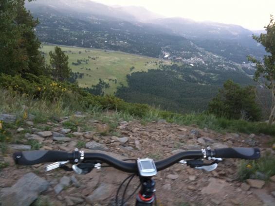 A view from the handlebars of a mountain bike looking down a rocky trail, with a vast landscape visible in the background, including hills, fields, and a distant town. The scene is captured at dusk, highlighting the natural beauty of the surrounding area. Bergen Peak mountain bike trail.