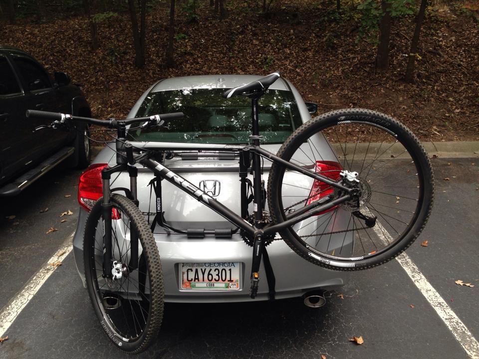 Trek X-Caliber: A black mountain bike secured on a bike rack attached to the back of a silver car parked in a wooded area. The car's rear plate is visible, and the surrounding ground is covered with autumn leaves.