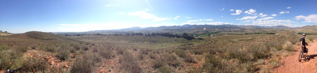 A panoramic view of a rugged landscape showcasing rolling hills and distant mountains under a clear blue sky. In the foreground, a dirt path bordered by sparse vegetation leads to a cyclist standing on the right, enjoying the expansive scenery. The composition captures the natural beauty and tranquility of the outdoors. Devil's Backbone mountain bike trail.