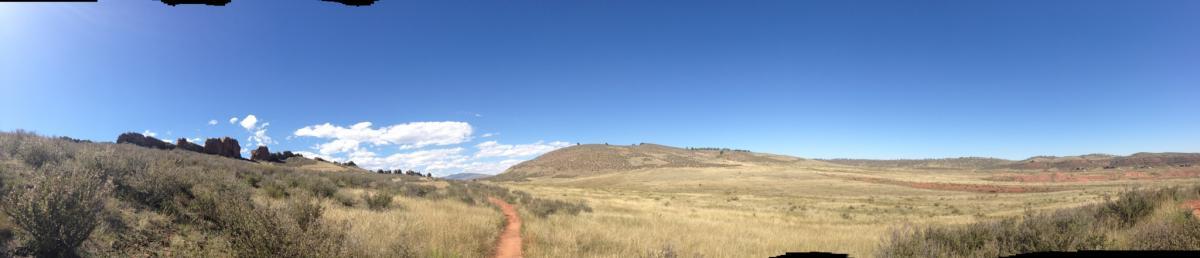 A panoramic view of a grassy landscape under a clear blue sky, featuring a winding dirt path leading into the distance. Low hills and rocky formations are visible on the left, with patches of greenery scattered throughout the scene. Devil's Backbone mountain bike trail.
