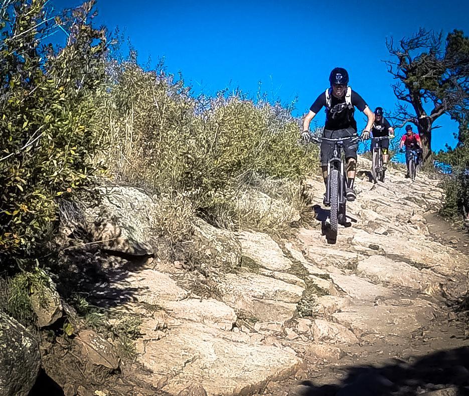 A group of mountain bikers navigating a rocky trail surrounded by greenery under a clear blue sky. Red Rocks / Dakota Ridge mountain bike trail.