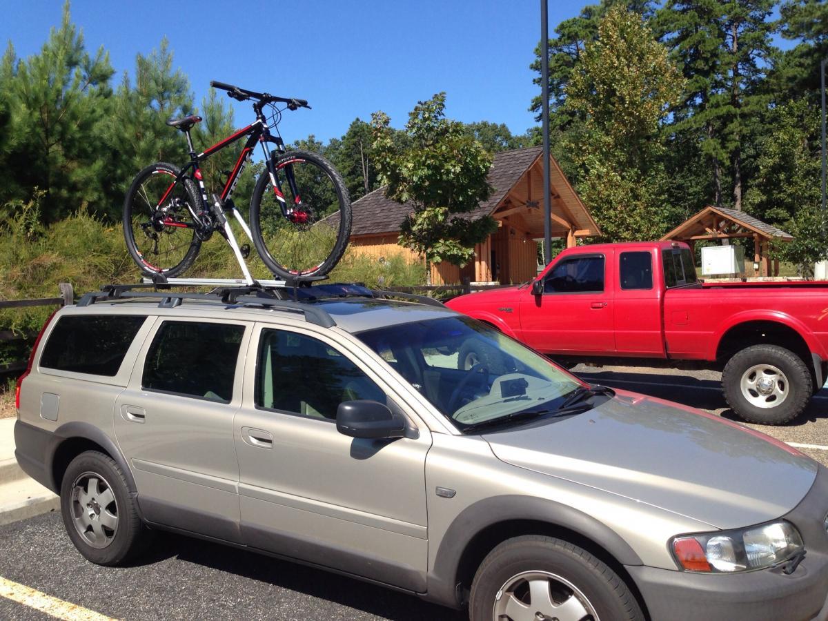 Trek Marlin: A silver SUV parked in a lot with a red mountain bike secured on its roof rack. In the background, there is a red pickup truck and trees, with a building partially visible. The scene is set under a clear blue sky.