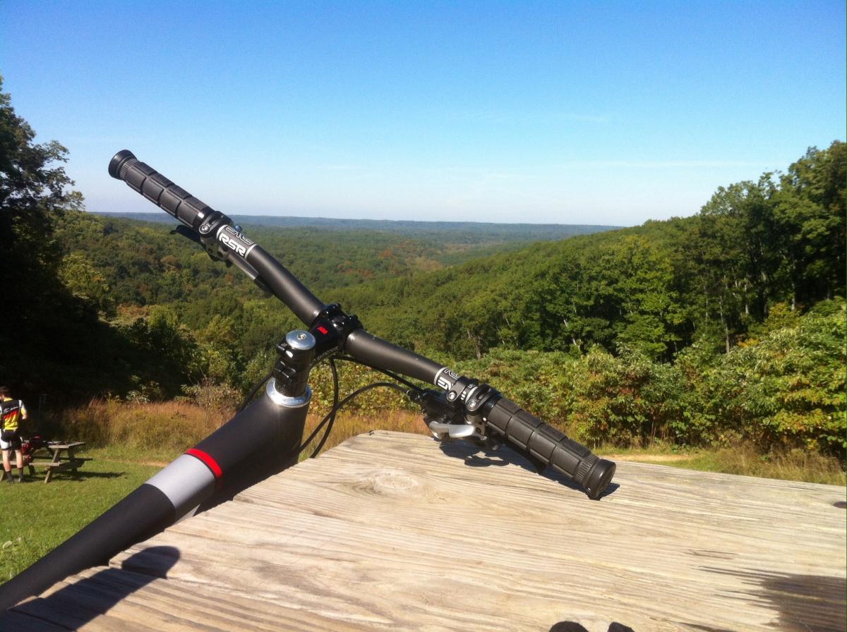 Close-up view of a mountain bike's handlebars resting on a wooden table, overlooking a lush green valley under a clear blue sky. In the background, rolling hills and trees create a scenic landscape, while a person with a bike is visible in the distance. Brown County Park mountain bike trail.