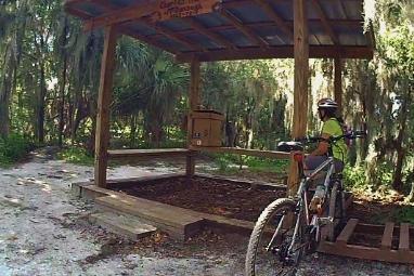 A mountain biker in a bright yellow shirt is resting next to a wooden shelter in a wooded area. The shelter has a sign and a wooden bench, surrounded by lush greenery and moss-covered trees. A mountain bike is parked nearby on the gravel path. Loyce E. Harpe Park mountain bike trail.