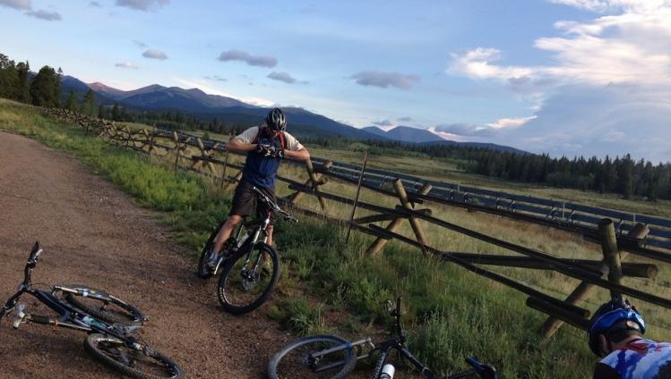 A scenic outdoor landscape featuring two mountain bikers resting on a gravel path surrounded by tall grass and a wooden fence. In the background, a range of mountains is visible under a partly cloudy sky. Several bicycles are laid on the ground nearby. Colorado Trail: Kenosha Pass To Breckenridge mountain bike trail.