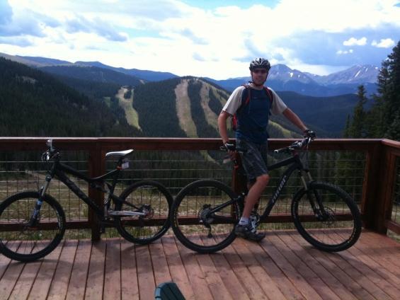 A mountain biker stands next to two bicycles on a wooden deck overlooking a scenic mountainous landscape with green forests and distant peaks under a partly cloudy sky. The biker is wearing a helmet and casual outdoor clothing. Keystone Resort Bike Park mountain bike trail.