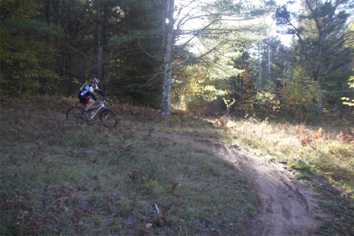 A mountain biker navigating a curved dirt trail through a wooded area with trees and autumn foliage in the background. Makwa Trail mountain bike trail.