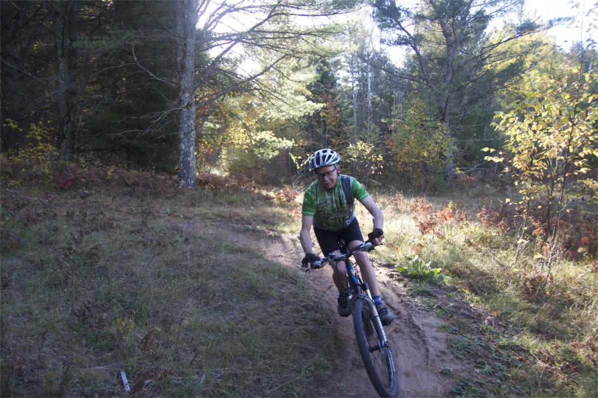 A mountain biker riding along a dirt trail in a forest during autumn, surrounded by trees and colorful foliage. The cyclist is wearing a green jersey and a helmet, showcasing an active outdoor lifestyle. Makwa Trail mountain bike trail.