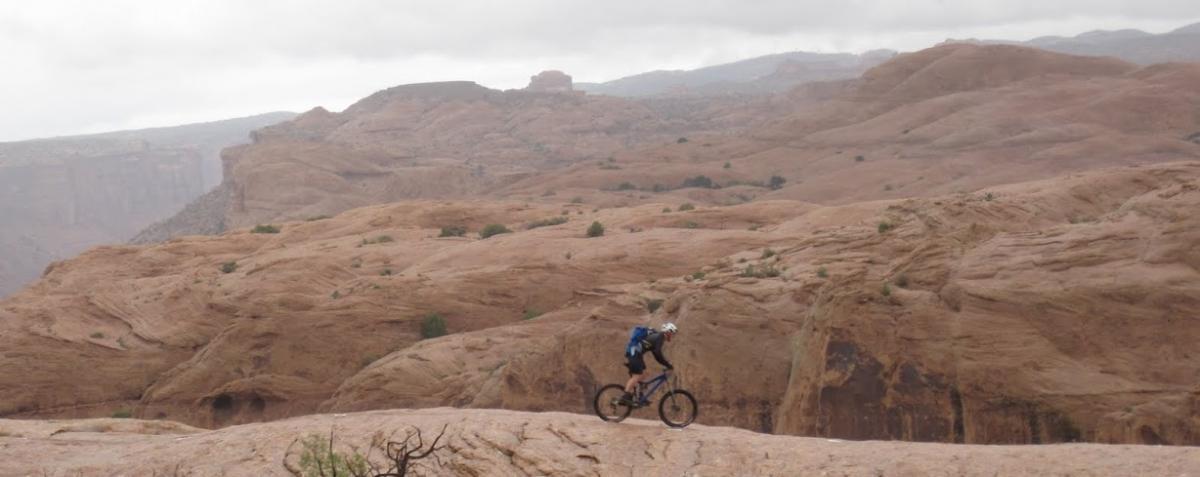 A mountain biker rides along a rocky terrain with rolling hills and dramatic cliffs in the background under a cloudy sky. Slickrock mountain bike trail.