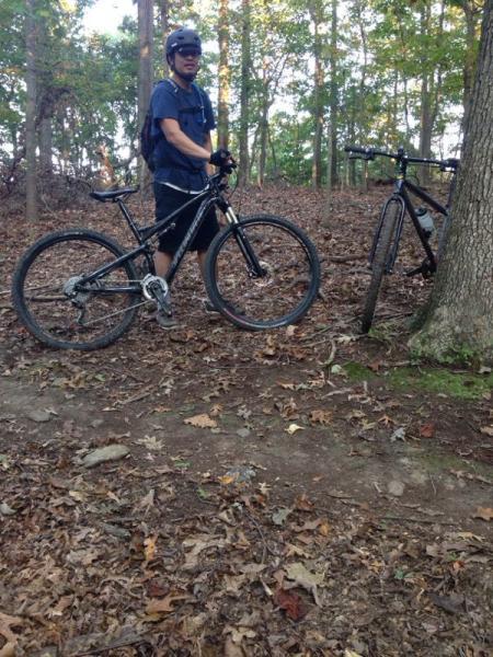 A person wearing a helmet and casual clothing stands next to a mountain bike in a wooded area, with another bike leaning against a tree. The ground is covered in fallen leaves, and the background features trees and natural foliage. Ronald C. McDonnell Campground Loop mountain bike trail.