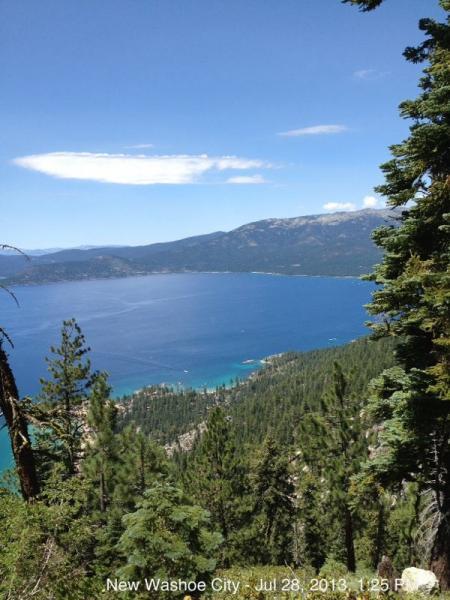A scenic view of Lake Tahoe from New Washoe City, showcasing vibrant blue water surrounded by lush green forests and distant mountains under a clear blue sky. Flume Trail mountain bike trail.