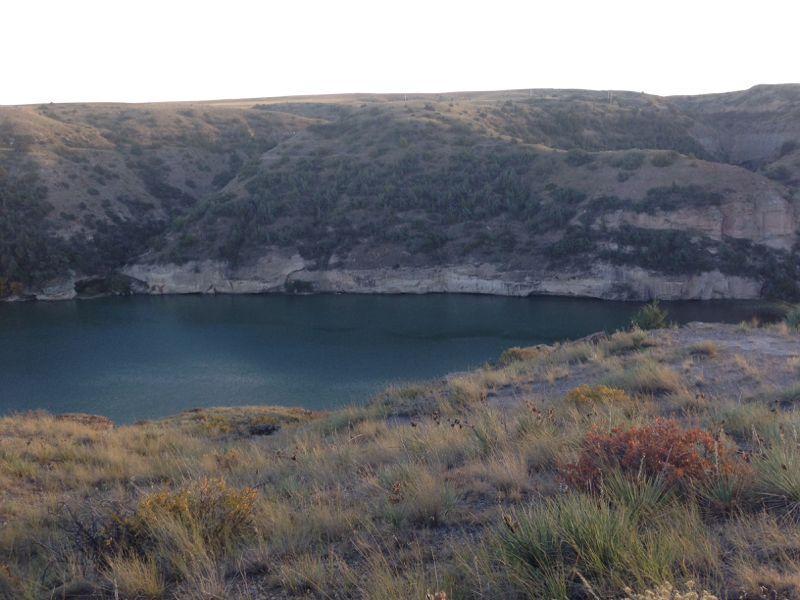 A serene landscape featuring a calm lake surrounded by rolling hills and greenery, with the sun setting in the background. The water reflects the soft hues of the sky, creating a tranquil scene in nature. North Shore mountain bike trail.