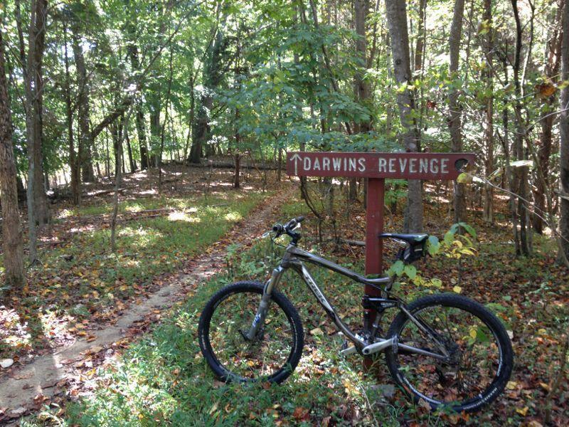Mountain bike parked beside a sign that reads "Darwin's Revenge," amidst a wooded trail surrounded by green trees and fallen leaves. Warriors' Path State Park mountain bike trail.