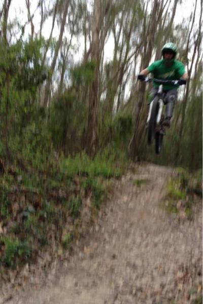 A person in a green shirt and helmet performing a jump on a mountain bike along a dirt trail, surrounded by trees and foliage. The image is slightly blurred, capturing the motion of the jump. Comite Trails mountain bike trail.