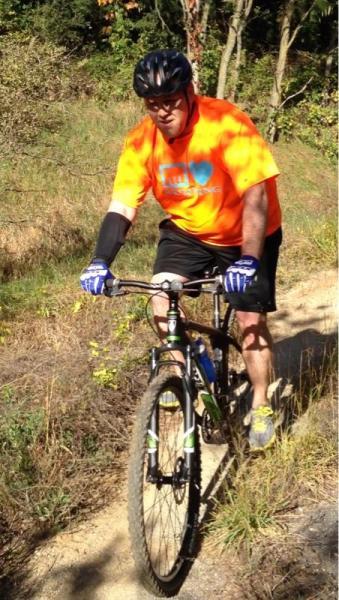 A person riding a mountain bike on a dirt trail surrounded by greenery. They are wearing an orange t-shirt, black shorts, and a cycling helmet, with gloves for added grip. The scene captures the individual in motion, navigating through a natural setting. Fort Custer Recreation Area mountain bike trail.