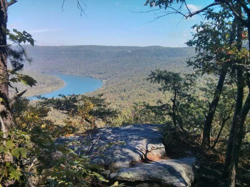 A panoramic view of a river winding through a lush green forest, captured from a rocky overlook. The scene features a clear blue sky with a few clouds, and trees framing the image in the foreground, highlighting the natural beauty of the landscape. Raccoon Mountain Trail Network mountain bike trail.