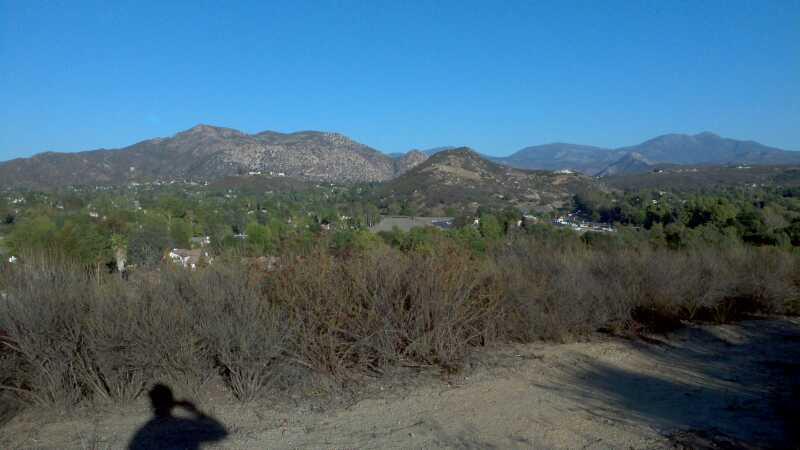 A scenic landscape featuring rolling hills and mountains under a clear blue sky. In the foreground, dry brush and sparse vegetation are visible, while the background showcases a green valley with small houses nestled among trees, bordered by distant mountains. A shadow of a person is cast in the lower left corner of the image. The Rollercoaster mountain bike trail.