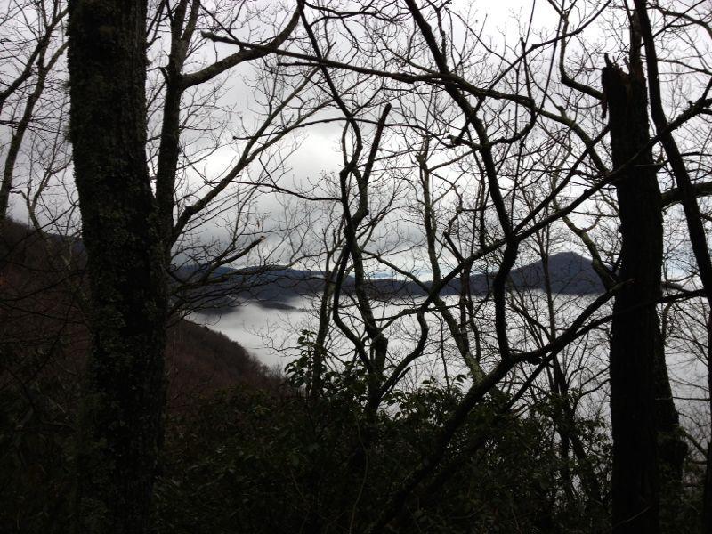 Alt text: A view of a misty landscape framed by bare tree branches, showcasing a serene body of water and distant mountains under a cloudy sky. Hickory Nut mountain bike trail.