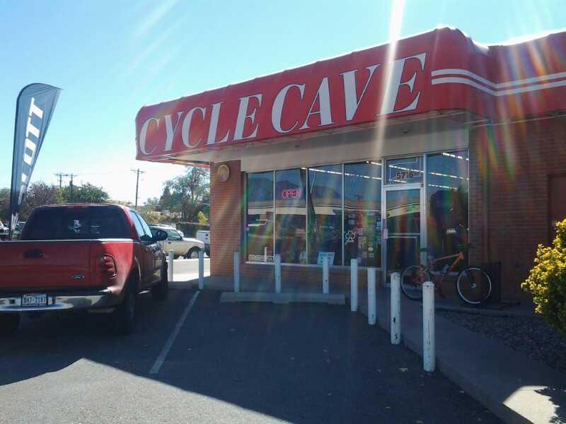 A storefront labeled "Cycle Cave" with a bright red awning. The shop displays an "Open" sign and features bicycles in the window. A red pickup truck is parked nearby, with a small flag on a pole to the left. The scene is set on a clear day, showcasing the shop's inviting atmosphere.