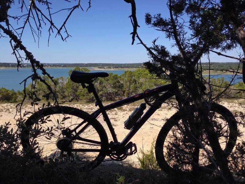 A mountain bike is silhouetted against a scenic view of a lake, framed by trees and bushes. The sky is clear with a few clouds, and the surrounding landscape features greenery and distant hills. Madrone Trail mountain bike trail.