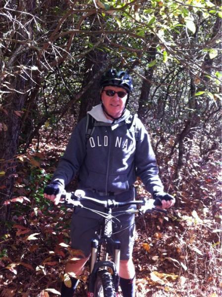 A person wearing a helmet and sunglasses stands next to a mountain bike in a wooded area, surrounded by trees and fallen leaves. They are dressed in a sweatshirt and shorts, ready for a biking adventure. Panthertown Valley mountain bike trail.