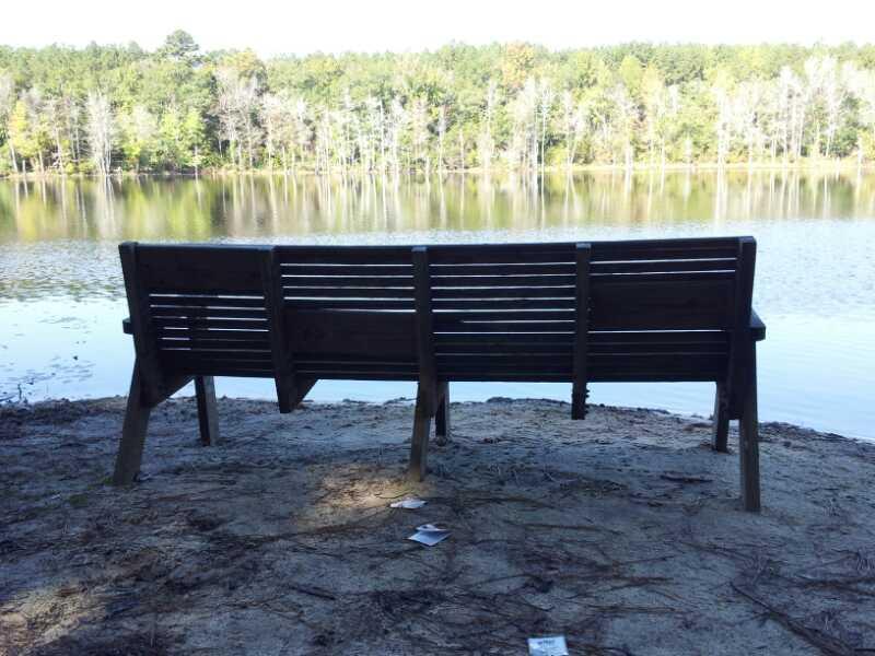 A wooden bench overlooking a serene lake, surrounded by trees. The view captures reflections in the water and hints of nature's tranquility, with some scattered items on the sandy shore. Boyd Pond mountain bike trail.