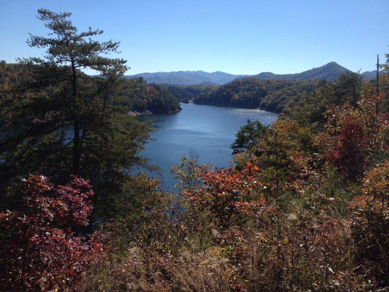 Scenic view of a serene lake surrounded by autumn-colored trees and rolling mountains under a clear blue sky. Tsali Recreation Area mountain bike trail.