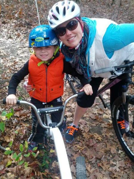 A young boy in a blue helmet and orange vest smiles while holding onto his bike, next to a woman wearing sunglasses and a colorful scarf. They are outdoors in a wooded area with fallen leaves, both appearing happy and engaged in a biking adventure. Needham Town Forest mountain bike trail.