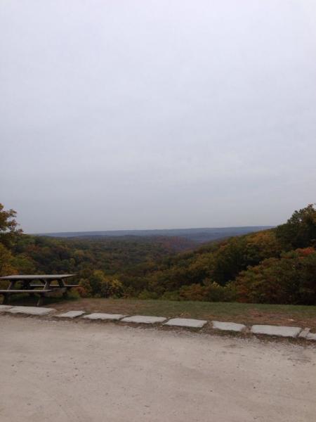 A panoramic view of a forested landscape under a cloudy sky, with rolling hills in the distance. In the foreground, a gravel path leads to a picnic table surrounded by trees displaying autumn colors. Brown County Park mountain bike trail.